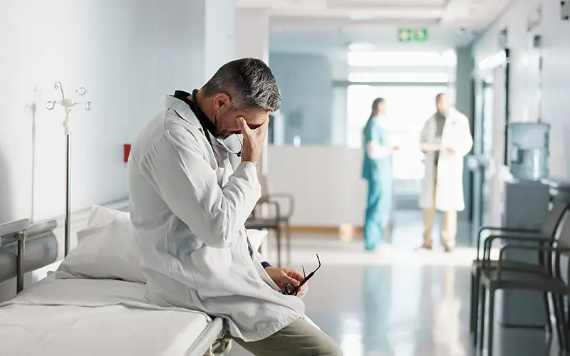 A physician in a white coat sitting on a gurney holding their head